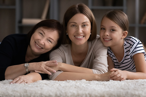 Those closest. Portrait of happy young woman mom daughter lying on floor while old age mommy and preteen child girl hug her from both sides. Loving family dynasty of 3 different females look at camera