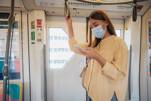 Asian business woman passenger wearing protective face mask using mobile phone and standing inside BTS skytrain or MRT subway at railway platform while traveling to work, Bangkok, Thailand, Transportation and lifestyle concept