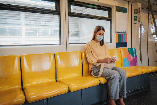 Asian business woman passenger wearing protective face mask using mobile phone and sitting inside BTS skytrain or MRT subway at railway platform while traveling to work, Bangkok, Thailand, Transportation and lifestyle concept