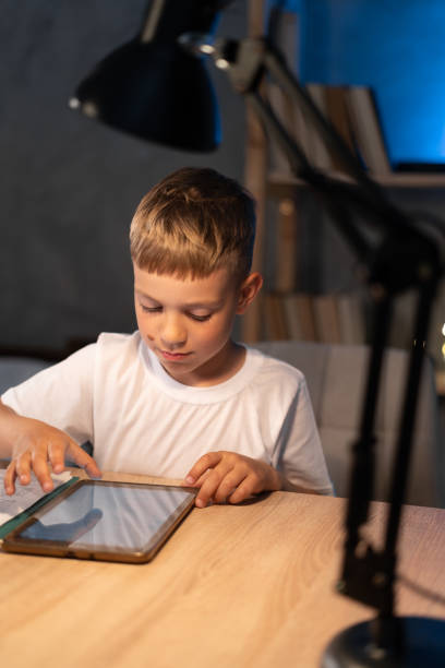 istock Boy looking at digital tablet while sitting at table at night, child reading e-book or watching cartoons using tablet 1410231434