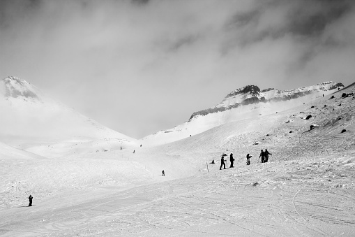 Skiers and snowboarders descent on snowy ski slope and overcast misty sky at day with bad weather before blizzard. Caucasus Mountains in winter, Georgia, region Gudauri. Black and white