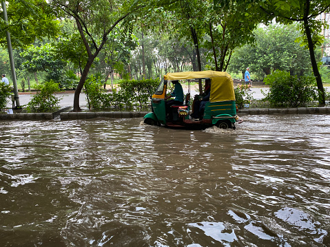 Ghaziabad, Uttar Pradesh, India - July 20, 2022: Stock photo showing traffic on a flooded road in a residential area, green and yellow auto rickshaw tuk tuks driving through waterlogged land caused by monsoon season.