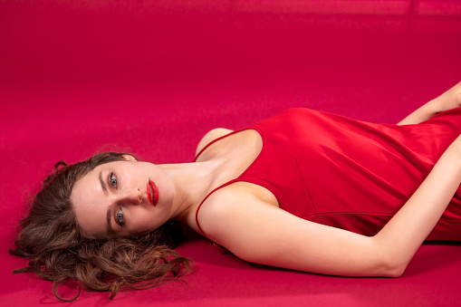 Close up portrait of a young woman with long brown hair in a red dress on a red background. She is lying on back and looking at camera.