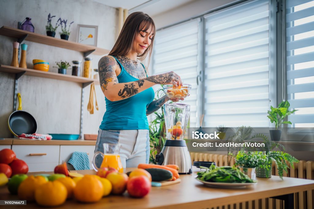 Woman making a healthy smoothie with fresh fruit Young smiling tattooed woman making a healthy smoothie with fresh fruit in her kitchen Adult Stock Photo
