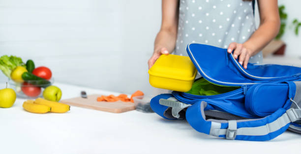 une mère attentionnée met une boîte à lunch en plastique jaune à son fils dans un sac à dos d’école. nourriture scolaire ou déjeuner, image concept. mise au point sélective, gros plan. - lunch box photos et images de collection