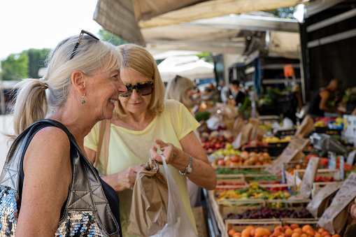 Two senior women out shopping for fruit at a farmer's market while on holiday in Tuscany, Italy. One woman is buying ingredients for sale over the counter and smiling.