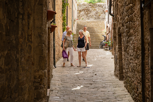 A senior couple and friends walking on a stone path through a street in Tuscany, Italy. They are exploring while talking with each other.