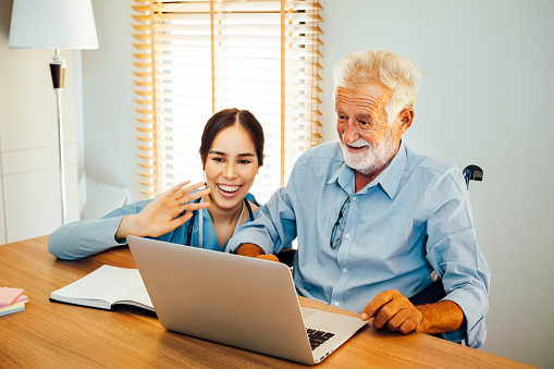 Smiling elderly businessman working with video conference and supervised by nurse.