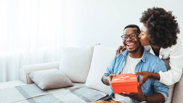 jovem casal com caixa de presente abraçando em casa. - men african descent giving flower - fotografias e filmes do acervo