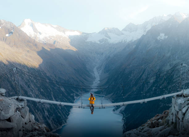 Man in yellow jacket sitting on a suspension bridge over a lake in Olpererhutte, Austria A mountainous landscape with a beautiful lake in the background in the Austrian alps a single person observes the amazing landscape and the immensity suspension bridge stock pictures, royalty-free photos & images