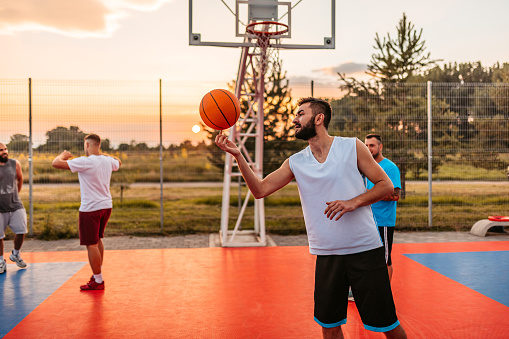 Young male basketball player standing on the basketball court with his friends and spinning a ball on his finger.