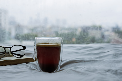 A glass of hot coffee with book and spectacles on bed in morning with rain drop on window. Stay home and relaxing concept.