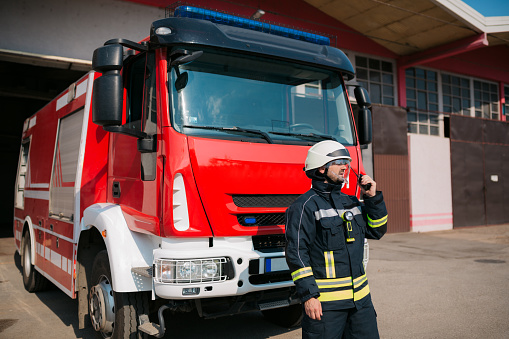 Firefighter standing in front of the fire truck, dressed in their uniform and using a radio