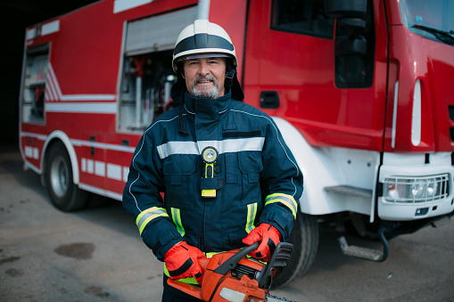 Portrait of a professional fireman standing in a fire station garage next to the fire truck, holding a chainsaw in his hands and looking at the camera