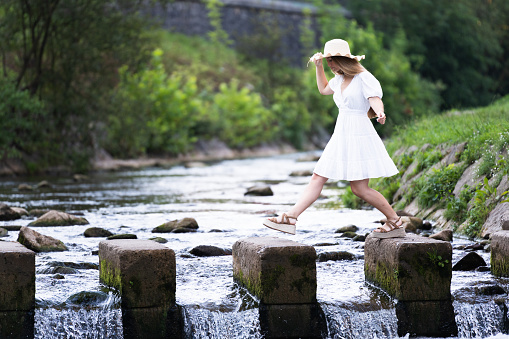 Beautiful teenager girl wearing white dress crossing river . Young tourist woman in nature