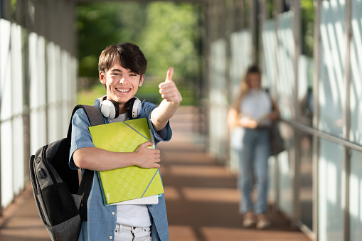 Cute student making ok sign and looking at camera. Portrait of positive boy in a corridor