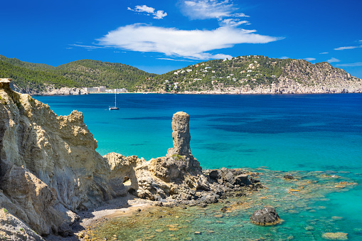 Wide-angle view of a long stretch of the north-eastern coastline of Ibiza, from the cliffs of Es Figueral to the promontory of Cala de Sant Vicent at the distance. The natural pillar known as Es Paller d'en Camp, separating the beach of S'Aigua Blanca from the beach of Es Figueral, is shown at the centre of the picture. Astonishingly transparent waters marbled by Posidonia meadows, the bright light of a summer afternoon, picturesque clouds, colourful cliffs covered with lush vegetation, a sailing ship moored in the bay. Developed from RAW.