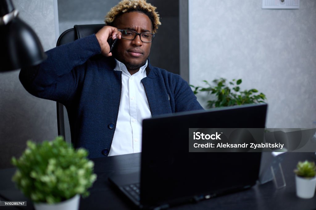 Successful employee of a company talking to a client feels confident African-American businessman who is engaged in sales using a smartphone, he has a busy working day in the office. Successful employee of a company talking to a client feels confident. Salesman Stock Photo