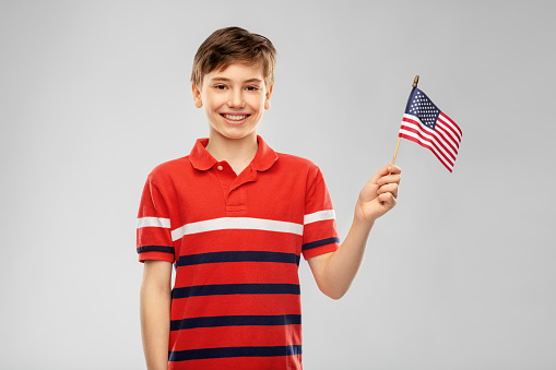independence day, patriotism and people concept - portrait of happy smiling boy in red polo t-shirt holding flag of united states of america over grey background