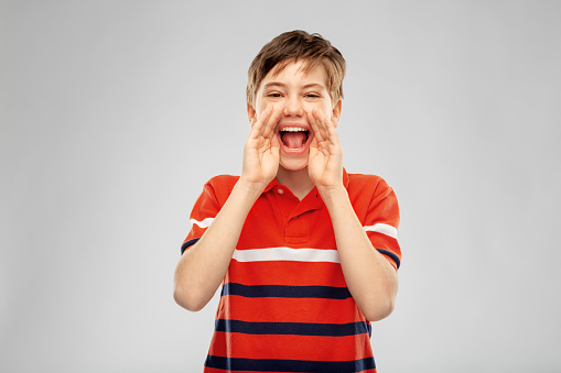 childhood, expressions and people concept - happy little boy in red polo t-shirt shouting or calling over grey background