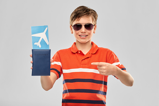 childhood, fashion and people concept - portrait of happy smiling boy in sunglasses and red polo t-shirt with air ticket and passport over grey background