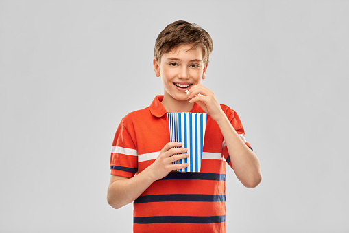 movie theater, cinema and people concept - portrait of happy smiling boy eating popcorn from striped bucket over grey background