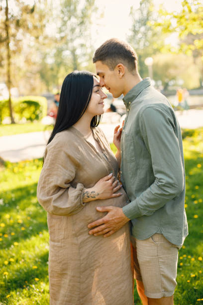 Pregnant woman and her husband standing in a park on a grass and hugging stock photo