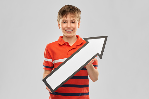 childhood, fashion and people concept - portrait of happy smiling boy in red polo t-shirt holding big white rightwards thick arrow over grey background over grey background