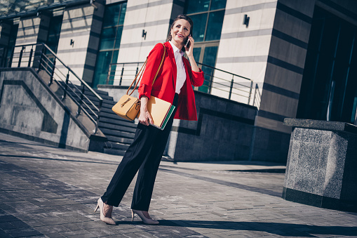 Photo of cheerful adorable mature lady secretary wear red jacket holding papers talking modern device outside city business complex.