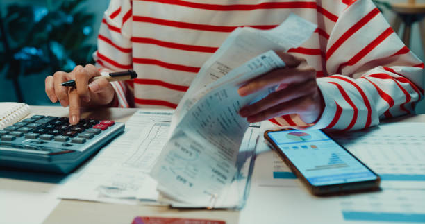 Close-up Young Asia woman with sweater sit in front of desk with phone use calculator to calculate utility bills check credit card receipt monthly expense bill in house at night. Home finance concept. primer plano mujer joven de asia con suéter se sienta frente al escritorio con calculadora de uso de teléfono para calcular las facturas de servicios públicos verifique la factura de gastos mensuales de recibo de tarjeta de crédito en la casa por la no