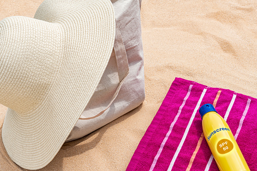 A straw hat sitting on a canvas tote bag that is on the beach sand next to a beach towel with a spray bottle of sunscreen.