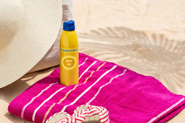 A straw hat sitting on a canvas tote bag that is on the beach sand next to a beach towel and flip flops with a spray bottle of sunscreen.