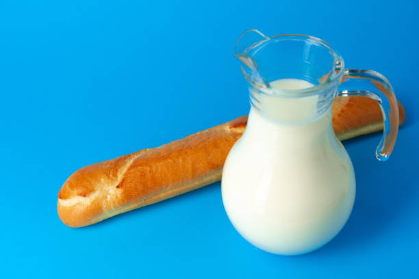 Fresh baguette bread and pitcher with fresh milk on blue background close up