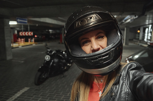Young beautiful woman in a helmet takes a selfie in a parking garage in front of her motorcycle.
