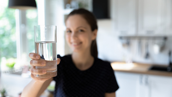 Happy young woman drinking clean mineral water in home kitchen, holding, showing transparent glass at camera, smiling. Close up beverage, banner shot. Healthy habit, hydration, lifestyle concept