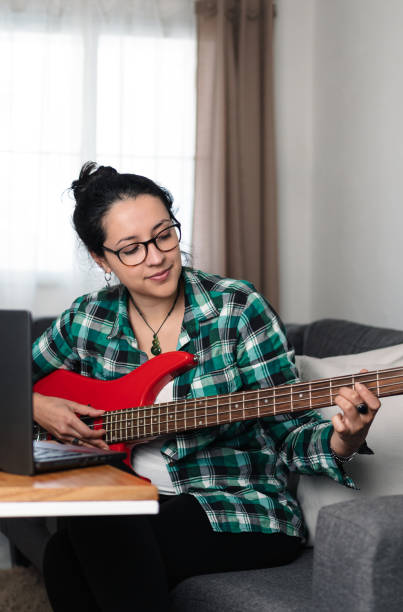 Brunette woman with glasses playing bass and using her computer at home stock photo