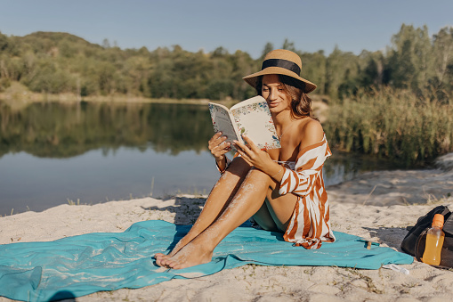 Beautiful brunette sitting on the beach towel and reading a book by the lake