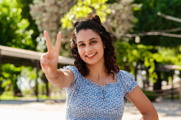 Young woman smiling confident wearing summer dress standing on city park, outdoors showing and pointing up with fingers number two while smiling confident and happy. Selective focus on her face. stock photo