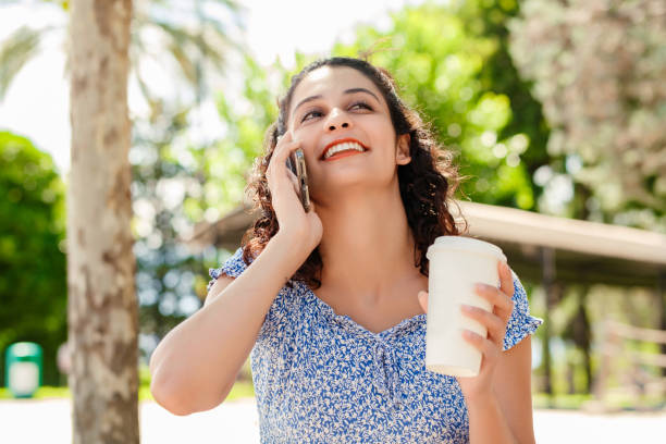 Young brunette girl smiling happy wearing summer dress on city park, outdoors talking phone and holding takeaway coffee mug while looking to sky. stock photo