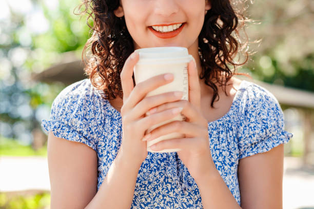 Young brunette girl smiling happy wearing summer dress on city park, outdoors holding a takeaway coffee mug front of chest. Coffee lover and outdoor concepts. stock photo