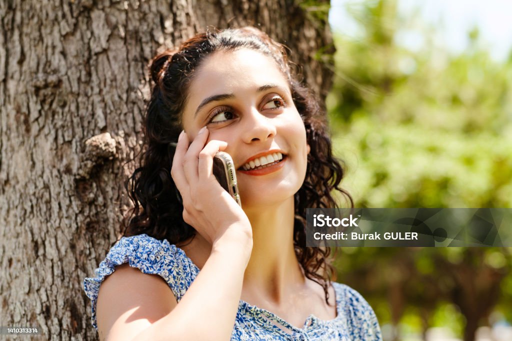 Young brunette girl smiling happy wearing summer dress on city park, outdoors talking on mobile phone with friends or boyfriend with smiles. Adults Only Stock Photo