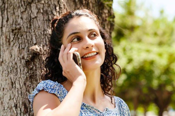 Young brunette girl smiling happy wearing summer dress on city park, outdoors talking on mobile phone with friends or boyfriend with smiles. stock photo
