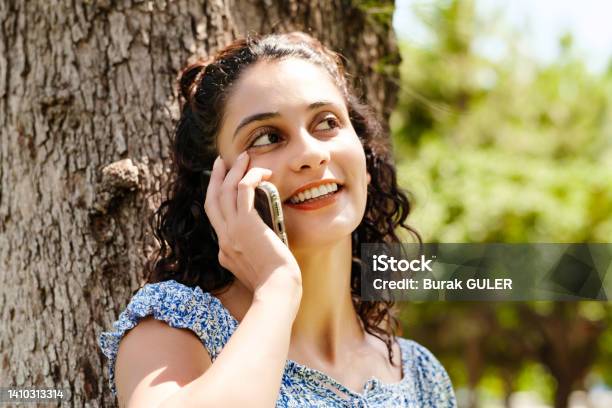 Young Brunette Girl Smiling Happy Wearing Summer Dress On City Park Outdoors Talking On Mobile Phone With Friends Or Boyfriend With Smiles Stock Photo - Download Image Now