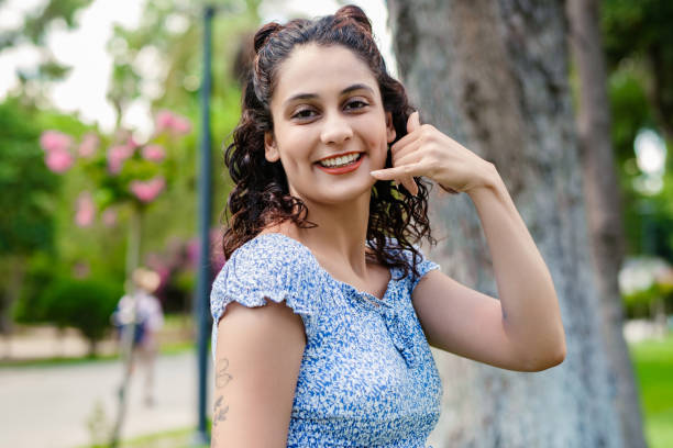 Young brunette girl smiling happy wearing summer dress on city park, outdoors makes a phone gesture and laughs looking at the camera. stock photo