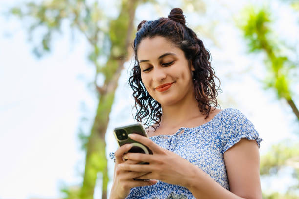Happy woman wearing summer dress standing on city park, outdoors looking at the phone screen and using phone. Messaging with friends, watching video or scrolling on social media. stock photo