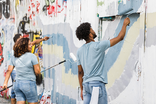 A mid adult mother and her young adult son cover graffiti with blue, purple, and grey waves of paint.