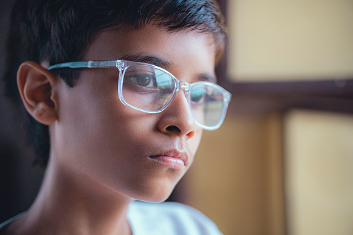 An Asian/Indian bespectacled teenage boy contemplates and looks at a view standing close to the window.