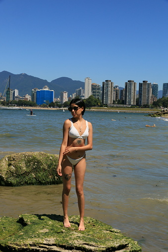 A Mexican woman standing on a rock in English Bay near Vancouver, BC. She is wearing a white bikini and sunglasses, necklace and tattoos.