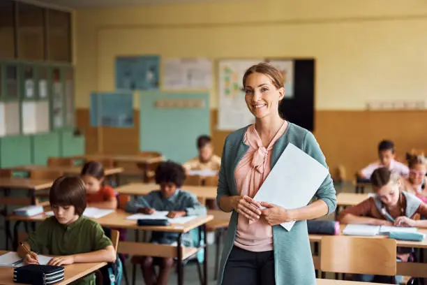 Happy teacher during a class at elementary school looking at camera. Her student are learning in the background.