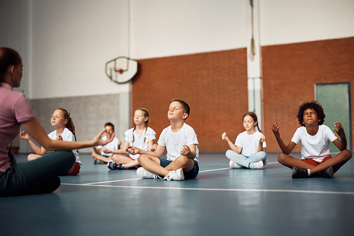 Elementary students meditating in lotus pose with their coach during PE class at school gym. Copy space.
